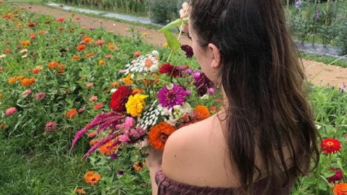 A person with long hair holds a colorful bouquet of flowers in a vibrant garden filled with blooming flowers.