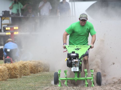 A man in a green shirt races a small tractor through dusty ground at a competition, with spectators in the background.