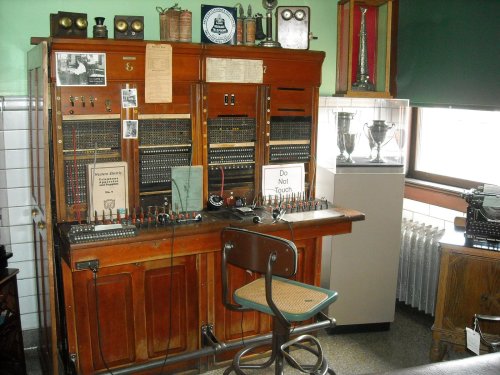 A vintage telephone switchboard with wooden panels, a chair, and various old communication equipment displayed.