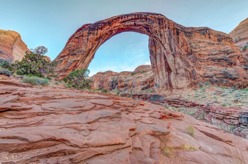 Natural rock arch surrounded by rugged red cliffs and sparse vegetation under a clear blue sky.