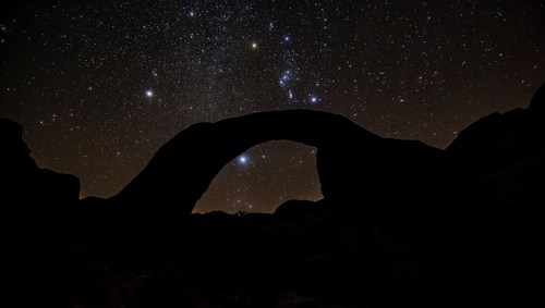 Silhouette of a natural arch against a starry night sky filled with bright stars and constellations.
