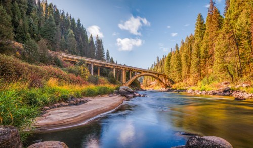 A serene river flows under a bridge, surrounded by lush green trees and a clear blue sky.