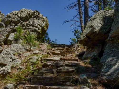 Stone steps lead upward between rocky formations under a clear blue sky, surrounded by sparse greenery.