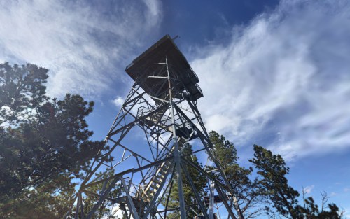 A tall metal fire lookout tower surrounded by trees under a blue sky with clouds.