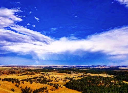 A panoramic view of rolling hills and fields under a bright blue sky with scattered clouds.