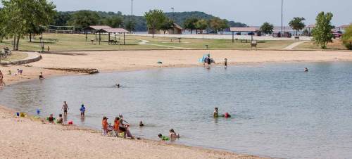 A sandy beach with people swimming and playing in the water, surrounded by trees and picnic areas.