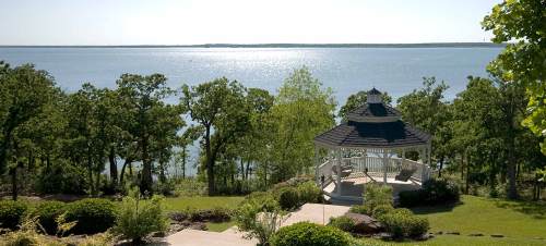 A scenic view of a gazebo overlooking a calm lake, surrounded by lush greenery and trees under a clear blue sky.