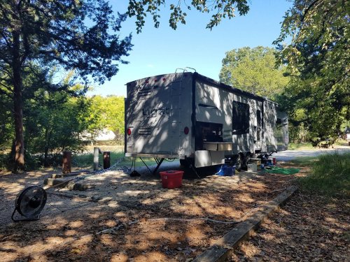 A parked RV surrounded by trees, with outdoor equipment and a clear blue sky in the background.