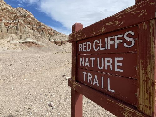 Sign for "Red Cliffs Nature Trail" in a desert landscape with rocky cliffs and blue sky in the background.