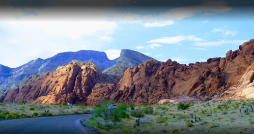 Scenic view of red rock formations and mountains under a blue sky, with a winding road in the foreground.