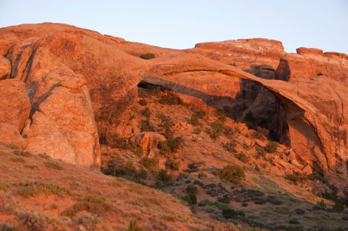 A natural stone arch rises above a rocky landscape, illuminated by warm sunlight during sunset.