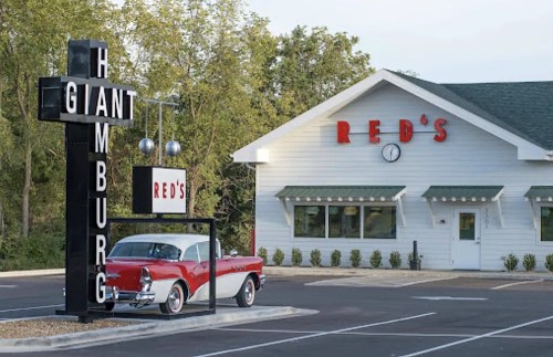 A vintage red car parked outside a diner named "Red's," featuring a large sign that reads "Giant Hamburg."