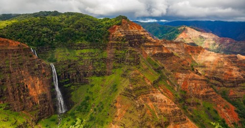 Aerial view of lush green mountains with a cascading waterfall and dramatic red rock formations under a cloudy sky.