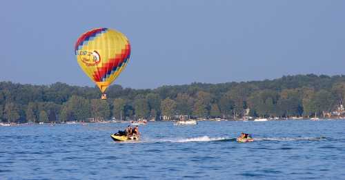 A colorful hot air balloon floats above a lake, while a jet ski tows a person on a tube nearby. Trees line the shore.