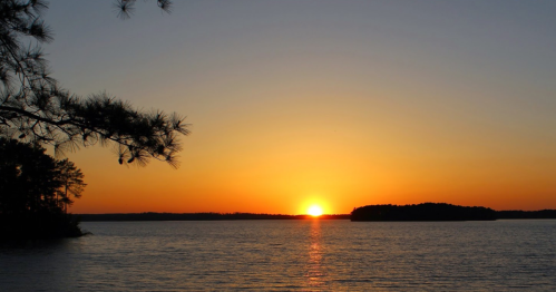 A serene sunset over a calm lake, with silhouettes of trees and islands against a vibrant orange sky.
