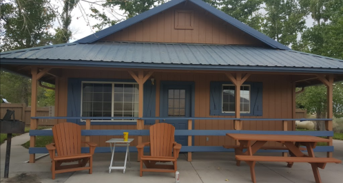 A cozy brown cabin with blue trim, featuring a porch, picnic table, and two wooden chairs in front.