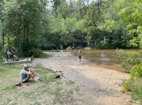 People enjoying a sunny day by a river, with some sitting on the shore and others playing in the water.
