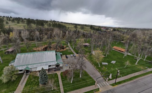 Aerial view of a park with green grass, trees, picnic areas, and a building under a cloudy sky.