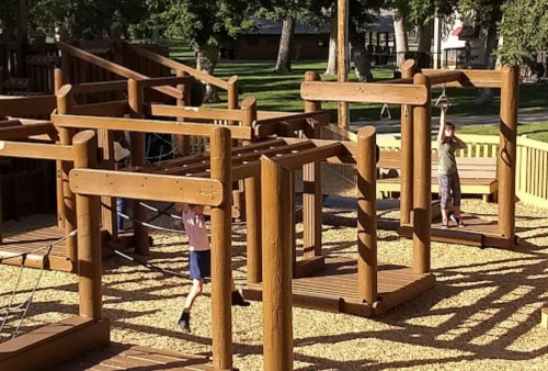 Children play on a wooden playground structure with swings and climbing elements in a sunny outdoor setting.