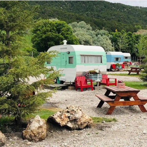 A vintage teal and white camper surrounded by greenery, with red chairs and picnic tables in a gravel area.