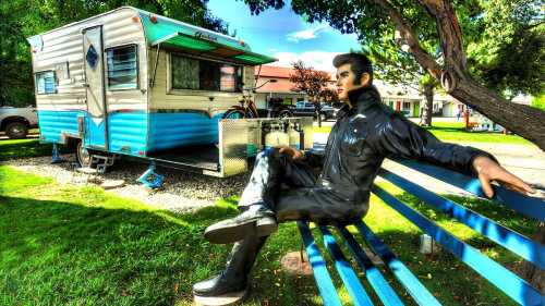 A statue of a man in a black outfit sits on a blue bench in front of a vintage camper trailer. Green trees surround the scene.