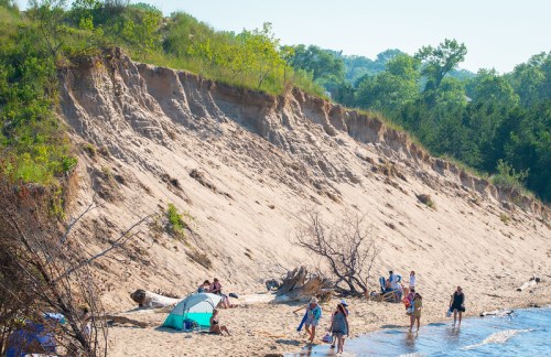 A sandy beach with a steep dune, people enjoying the sun, and a few tents near the water's edge.