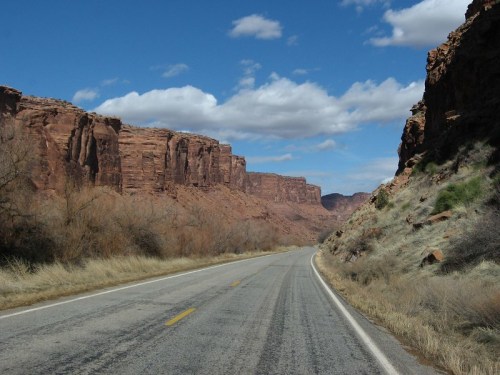 A winding road through a canyon with towering red rock formations and a blue sky dotted with clouds.