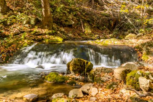 A serene stream flows over rocks, surrounded by autumn foliage and trees, creating a peaceful natural scene.