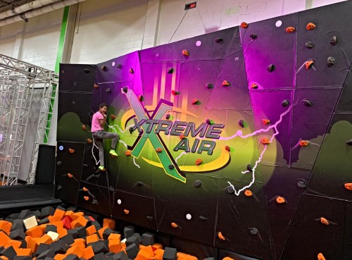 A child climbs a colorful rock wall at an indoor climbing facility, surrounded by foam blocks.