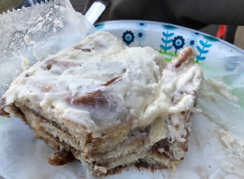 A slice of cinnamon roll cake with cream cheese frosting on a paper plate, partially eaten.