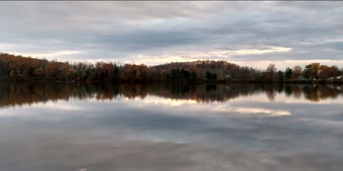 A calm lake reflects autumn trees and a cloudy sky, with a hint of sunset colors on the horizon.