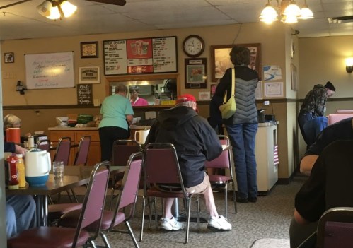 A cozy diner scene with patrons seated at tables, a counter with staff, and a menu board on the wall.