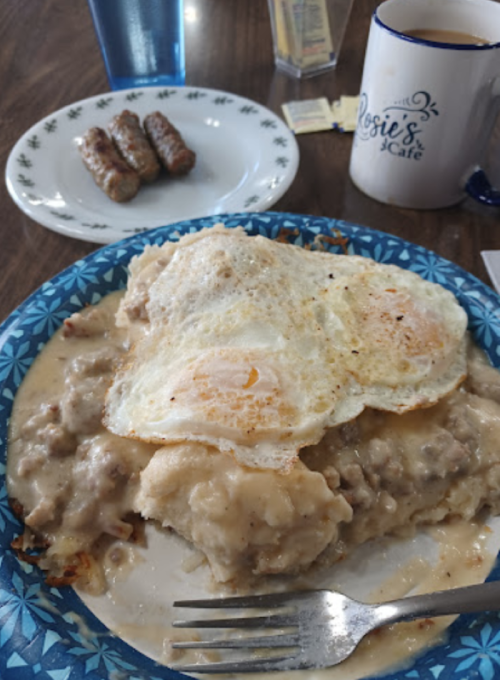 A plate of biscuits and gravy topped with two fried eggs, with a side of sausage links and a coffee cup in the background.