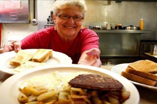 A smiling woman in a red shirt serves plates of breakfast food, including toast, eggs, and potatoes, in a diner setting.