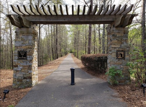 Stone archway marking the entrance to a wooded path in a park, with signs on either side and greenery surrounding the trail.