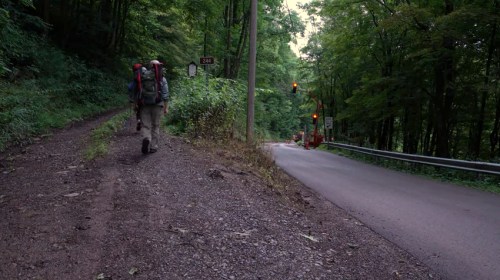 A hiker with a backpack walks along a dirt path next to a road with traffic lights and lush green trees.