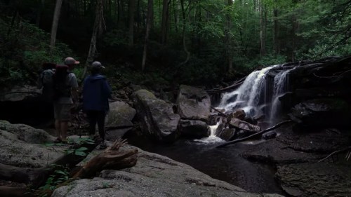Two hikers stand by a serene waterfall in a lush, green forest, surrounded by rocks and trees.