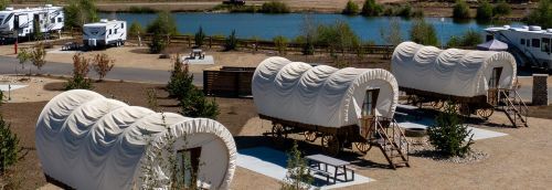 Three covered wagons are set up near a calm lake, surrounded by trees and picnic areas.