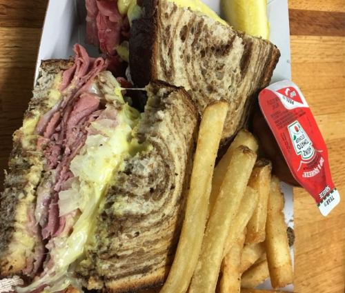 A close-up of a sandwich with layers of meat and cheese, served with fries and a small container of sauce.