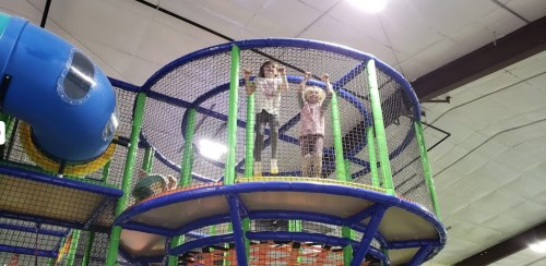 Two children play on a multi-level indoor playground, standing on a circular platform surrounded by safety netting.
