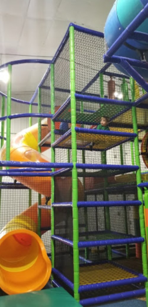 A colorful indoor play structure with slides and netting, featuring a child playing on the upper level.