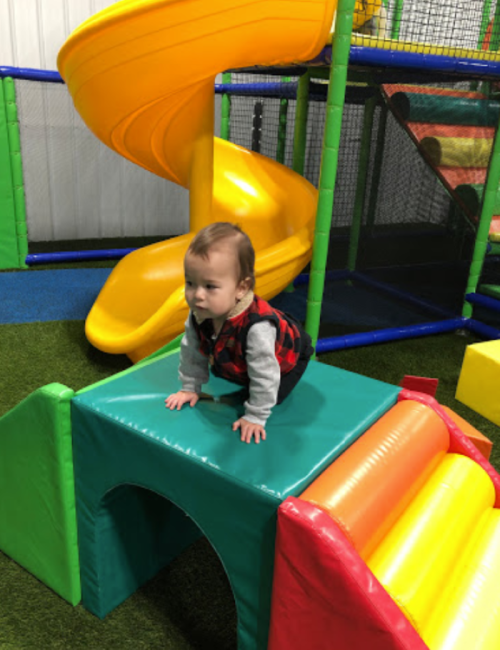 A toddler in a red and black vest sits on a colorful play structure in a playground with slides and soft surfaces.