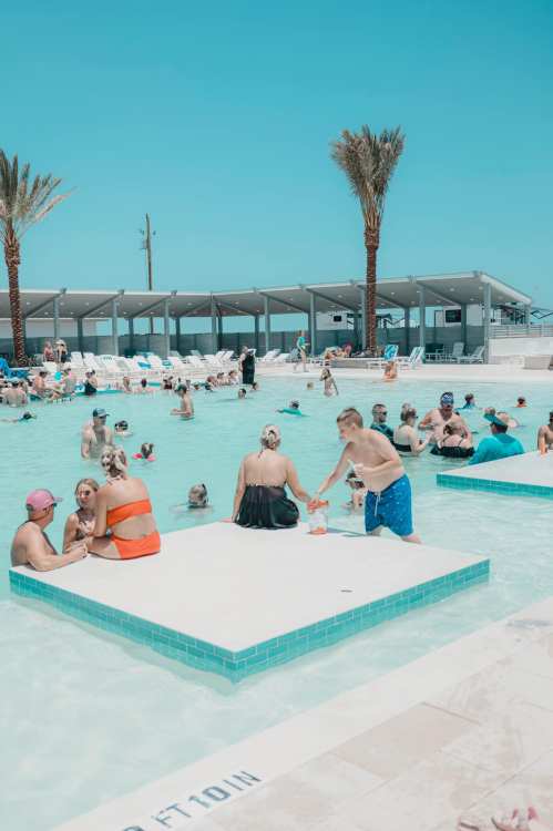 A lively pool scene with families and children enjoying the water under a clear blue sky, surrounded by palm trees.