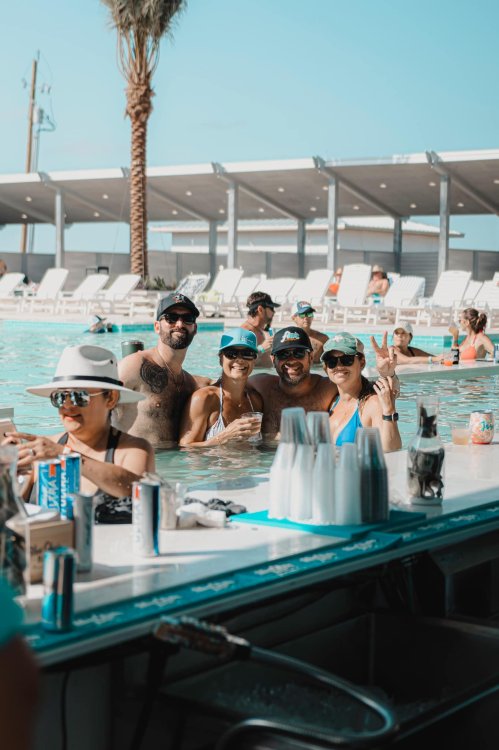 A group of four friends enjoying drinks in a pool, with lounge chairs and palm trees in the background.