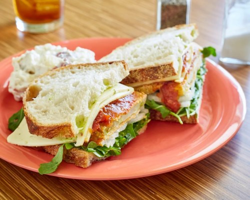 A close-up of a sandwich on a pink plate, filled with greens, cheese, and layers of meat, alongside a side dish.
