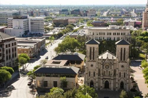 Aerial view of a cityscape featuring a historic church and modern buildings, with greenery and streets visible.