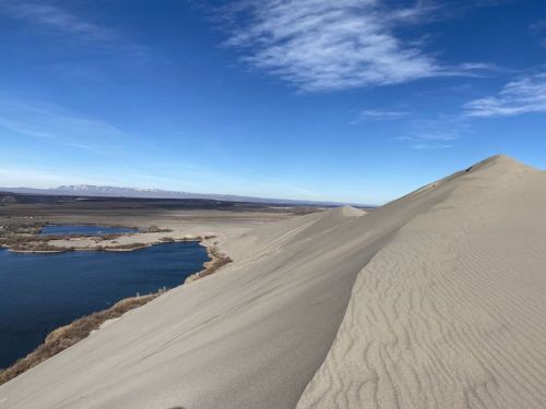 A sandy dune rises above a calm blue lake, with distant mountains under a clear blue sky.