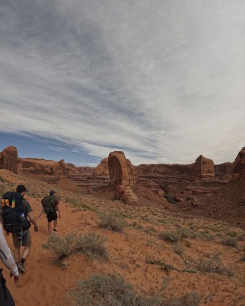 Hikers walking through a desert landscape with rock formations and a cloudy sky in the background.