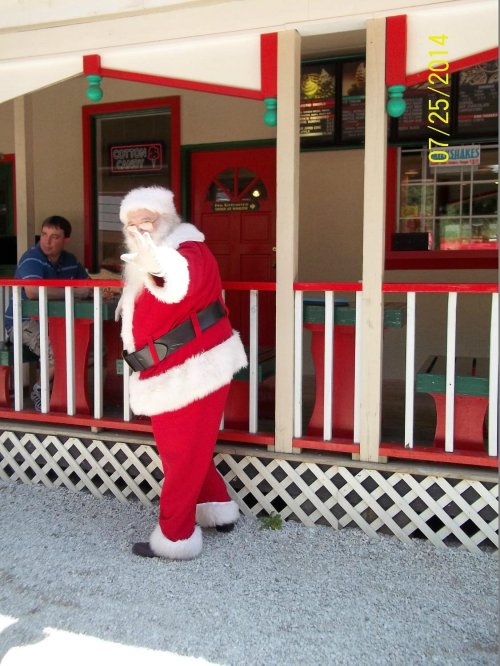 Santa Claus in a red suit waves from a colorful outdoor setting, with a person sitting nearby.