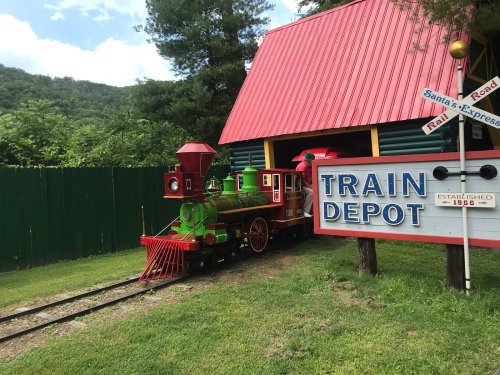 Colorful miniature train at a train depot with a sign, surrounded by green grass and trees under a blue sky.